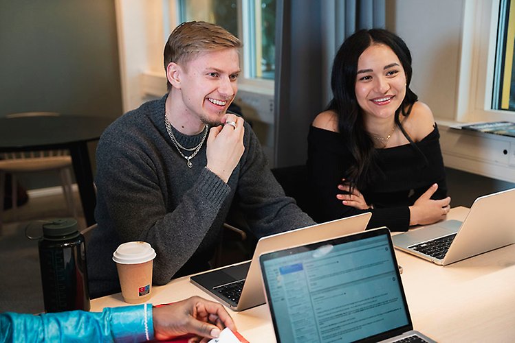 Bild på glada studenter som sitter och samtalar vid ett bord i orangeri med växter i bakgrunden. 