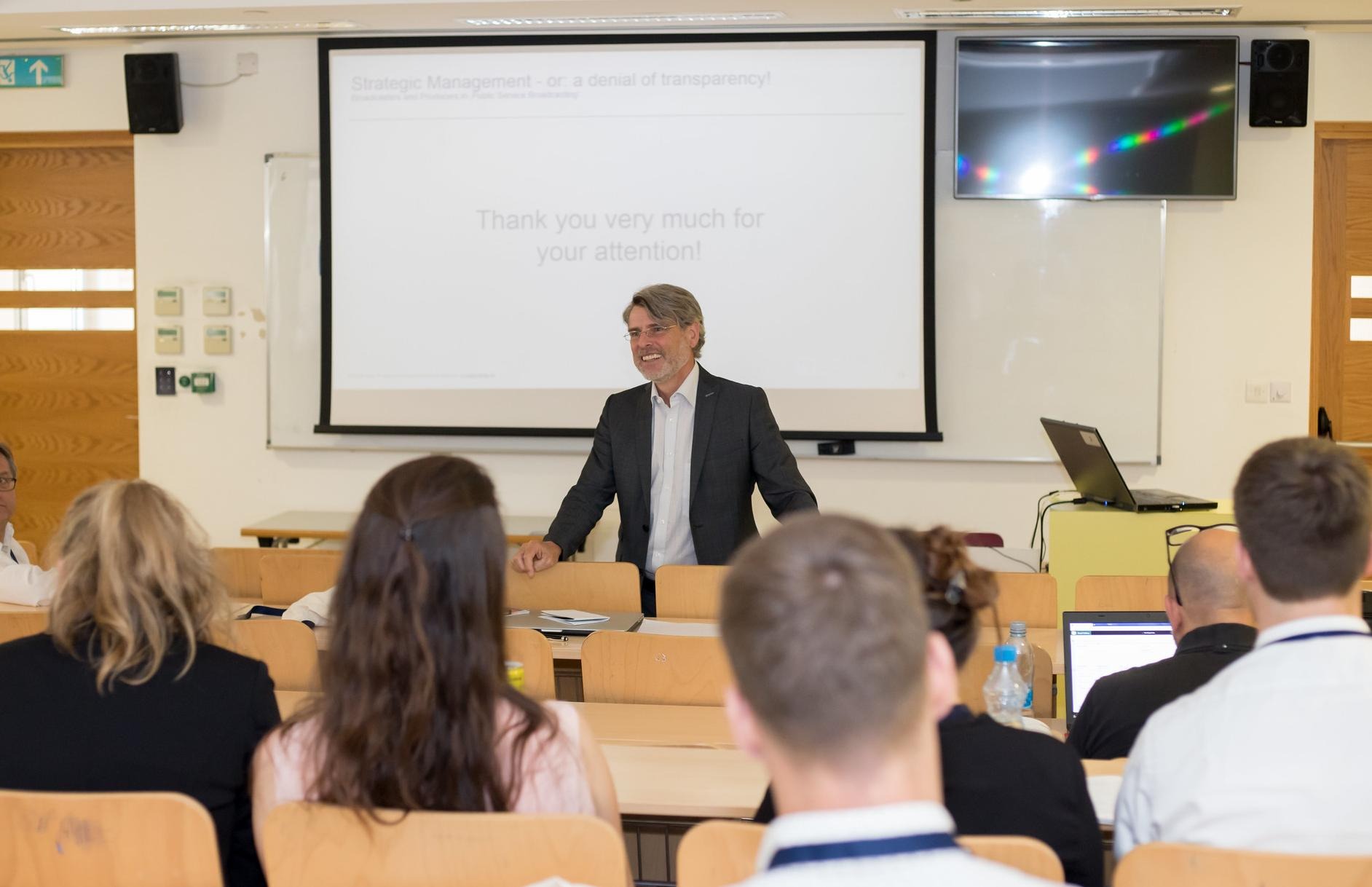 A male reseacher stands in front of a presentation screen in a lecture room. He is presenting to an audience who are sitting in rows with their backs to us.