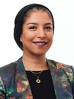 Woman wearing a dark headscarf, patterned blazer, and gold necklaces, smiling and facing forward against a white background.